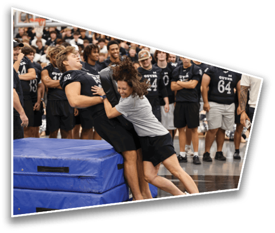 A person in a gray shirt playfully pushes another person in a black sports uniform against stacked blue gym mats while a group of people in matching black sports uniforms watch and smile in the background. Description generated by AI