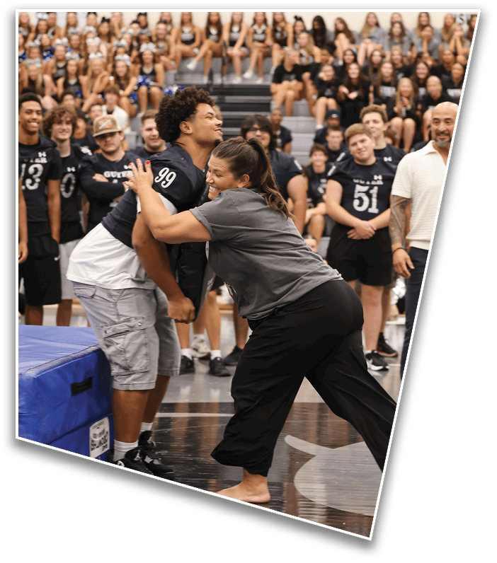 A person in casual clothing playfully pushes another person wearing a sports jersey in a gymnasium filled with people watching and smiling. Description generated by AI