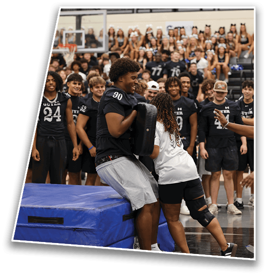 A group of people wearing sports jerseys are gathered in a gymnasium with one person holding a blocking pad while another person leans into it, surrounded by an audience in the background. Description generated by AI