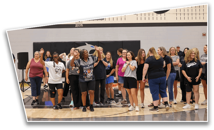 A group of adults standing and walking together in a gymnasium near a basketball hoop, some smiling and interacting with each other. Description generated by AI