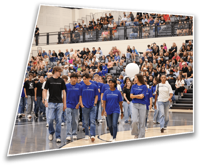 A group of people wearing matching blue shirts and jeans walk together on a gymnasium floor while a large crowd watches from the bleachers. Description generated by AI