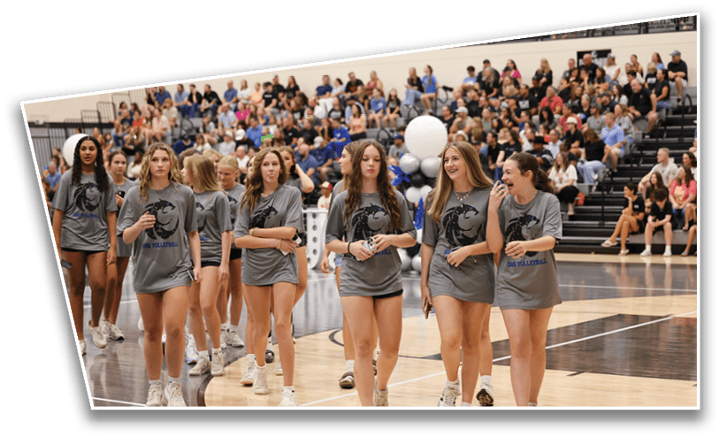A group of people wearing matching gray shirts and white shoes walk together on a gymnasium floor with a large crowd seated in the bleachers behind them. Description generated by AI