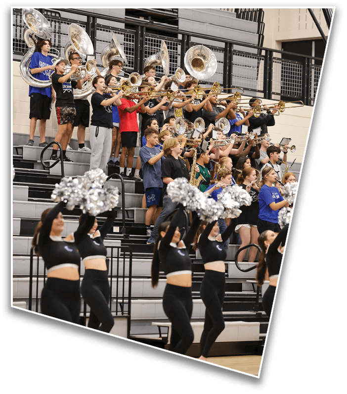 A group of people playing brass instruments in bleachers is accompanied by a line of people in black outfits holding silver pom-poms performing a cheer routine in a gymnasium. Description generated by AI