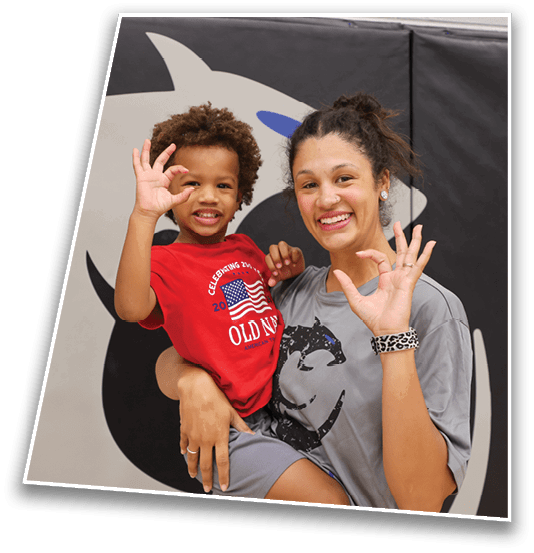 A person wearing a gray shirt is holding a child in a red shirt and gray shorts while both wave and smile against a background with black and beige colors. Description generated by AI