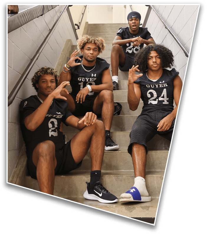 Four people wearing black sports uniforms with “GUYER" printed on them are sitting on a stairwell, with three seated on the steps and one higher up, all making hand gestures. Description generated by AI