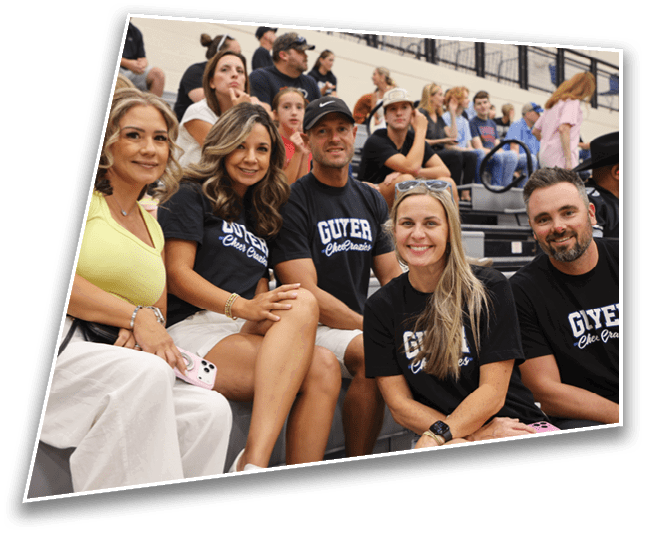A group of adults sitting closely together on bleachers, some wearing matching black shirts with white text, smiling and appearing engaged in an event. Description generated by AI