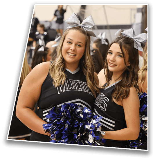 Two people wearing matching cheerleading uniforms with silver bows and holding blue and silver pom-poms smile while posing indoors. Description generated by AI