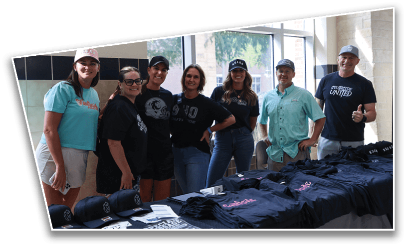 Seven adults wearing casual clothing and hats stand behind a table displaying neatly arranged folded t-shirts and caps in a bright indoor setting. Description generated by AI