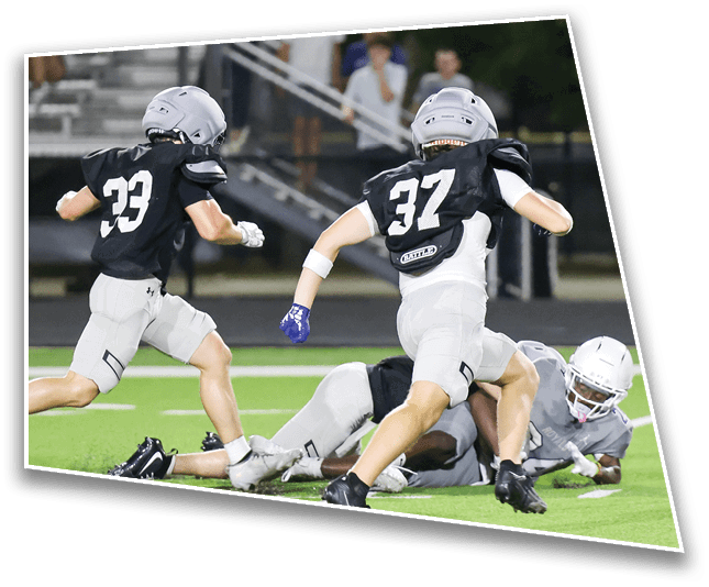 Two people wearing black jerseys and helmets are running on a football field while another person in gray gear is on the ground reaching for the ball. Description generated by AI