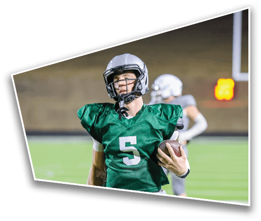 A person wearing a green football jersey with the number five is holding a football on a field with other players in the background. Description generated by AI