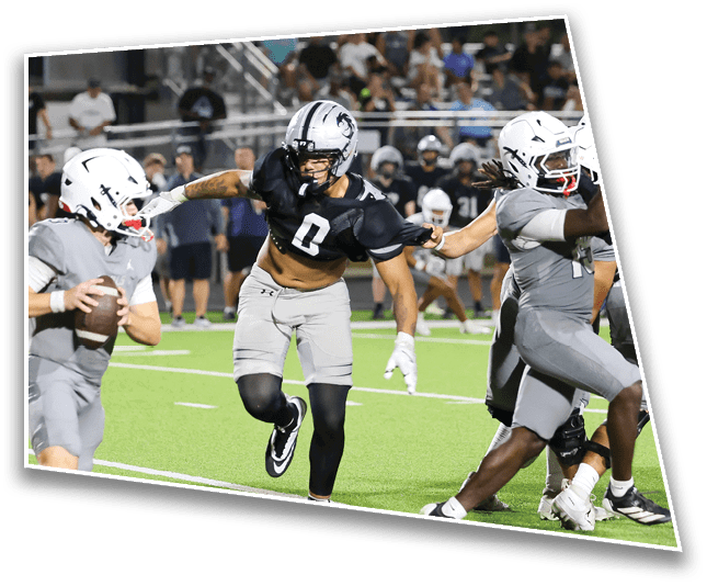 A person in a black sports jersey and helmet is reaching out to grab the arm of another person in a gray sports uniform on a football field during a game. Description generated by AI