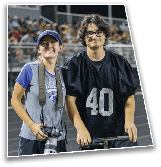 Two people stand side by side on a sports field, one holding a camera and wearing a cap and sports shirt, and the other wearing a black sports jersey with the number 40 and glasses. Description generated by AI
