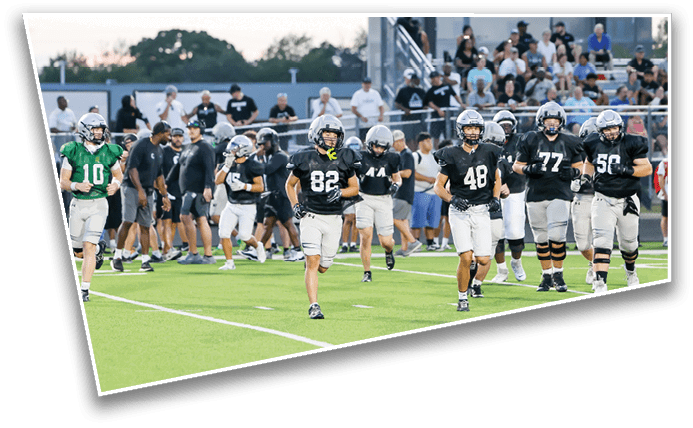 A group of football players in black and white uniforms are walking on a field with one player in a green jersey standing to the side and spectators watching from bleachers in the background. Description generated by AI