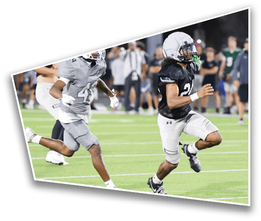 Two people in football uniforms are running on a field during a game with blurred spectators in the background. Description generated by AI