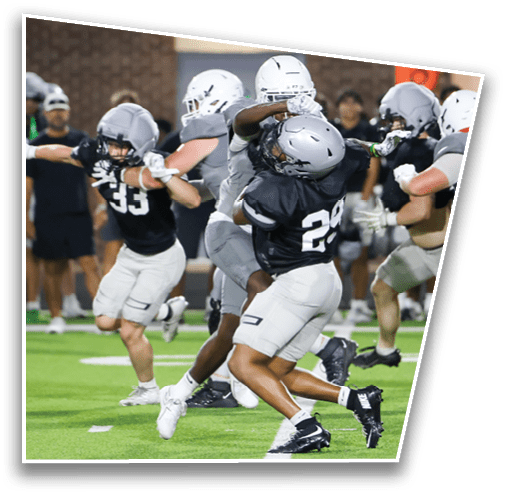 People wearing helmets and football uniforms are actively engaged in a tackle during a game on a green field with spectators in the background. Description generated by AI