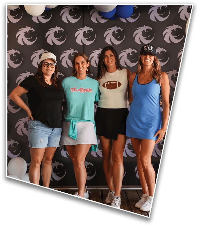 Four adults stand closely together smiling in front of a patterned backdrop with balloons, wearing casual summer clothing including hats and shorts or dresses. Description generated by AI