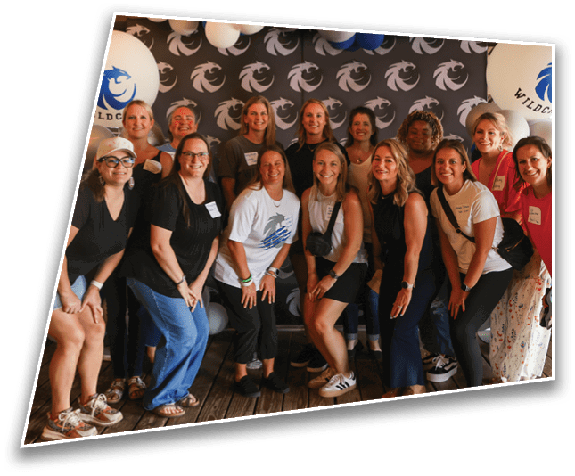 A group of smiling adults posing together indoors in casual clothing with a backdrop featuring a repeating logo and balloons. Description generated by AI