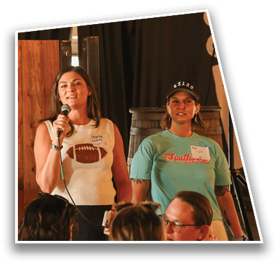 Two adults wearing casual clothing and name tags stand indoors near wooden barrels while other people sit in front of them. Description generated by AI