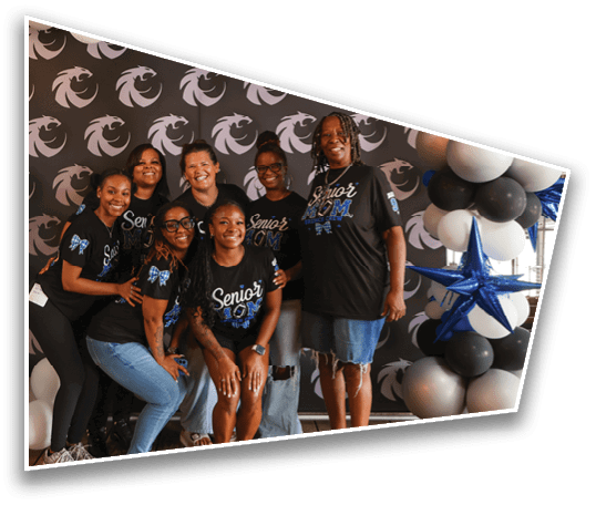 A group of seven people wearing matching black “Senior" shirts pose smiling in front of a backdrop with a repeating logo and a balloon arrangement with black, white, silver, and blue star-shaped balloons. Description generated by AI