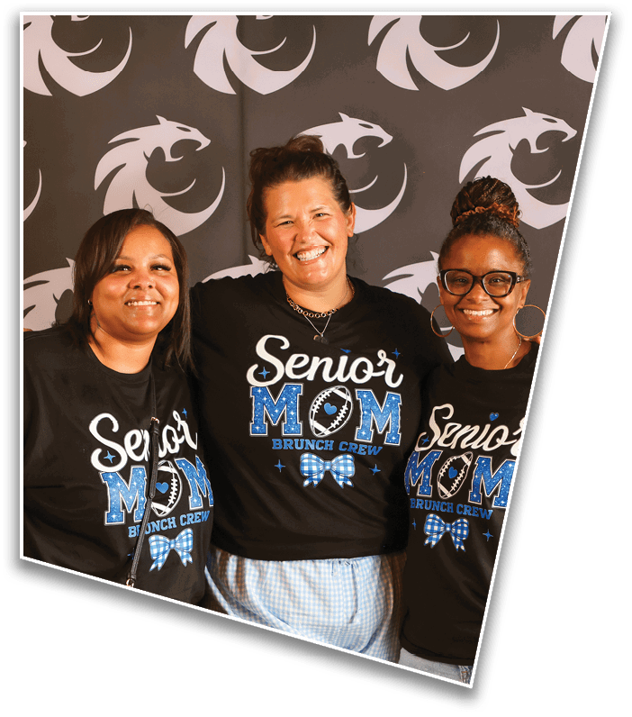 Three adults smiling and standing close together wearing matching black shirts that say “Senior Mom Brunch Crew" with a football and bow design against a backdrop with a repeating logo. Description generated by AI