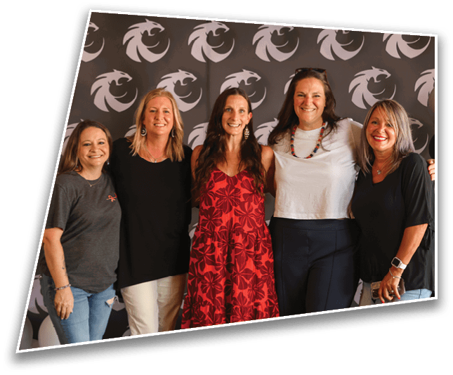 Five adults stand closely together smiling in front of a backdrop with a repeating dragon logo, wearing casual and semi-casual clothing. Description generated by AI