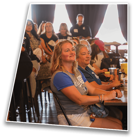 A group of people seated indoors with dark curtains and bright light coming from behind them. Description generated by AI