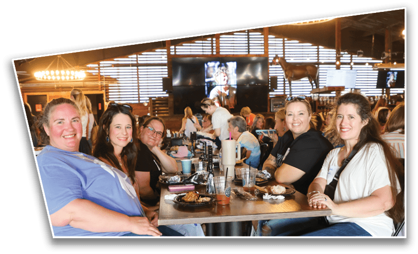 Five adults sitting around a restaurant table smiling with plates of food and drinks in front of them in a warmly lit casual dining setting. Description generated by AI