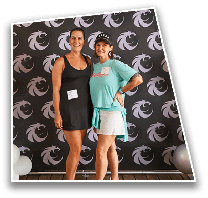 Two adults stand side by side on a wooden floor in front of a black backdrop with a repeating dragon logo, one wearing a black dress and flip-flops and the other wearing a turquoise shirt, white skirt, and white sneakers. Description generated by AI