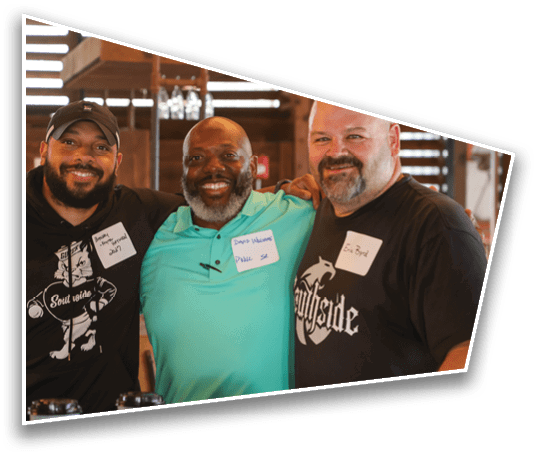 Three adults smiling and standing close together indoors with name tags on their shirts and casual clothing. Description generated by AI