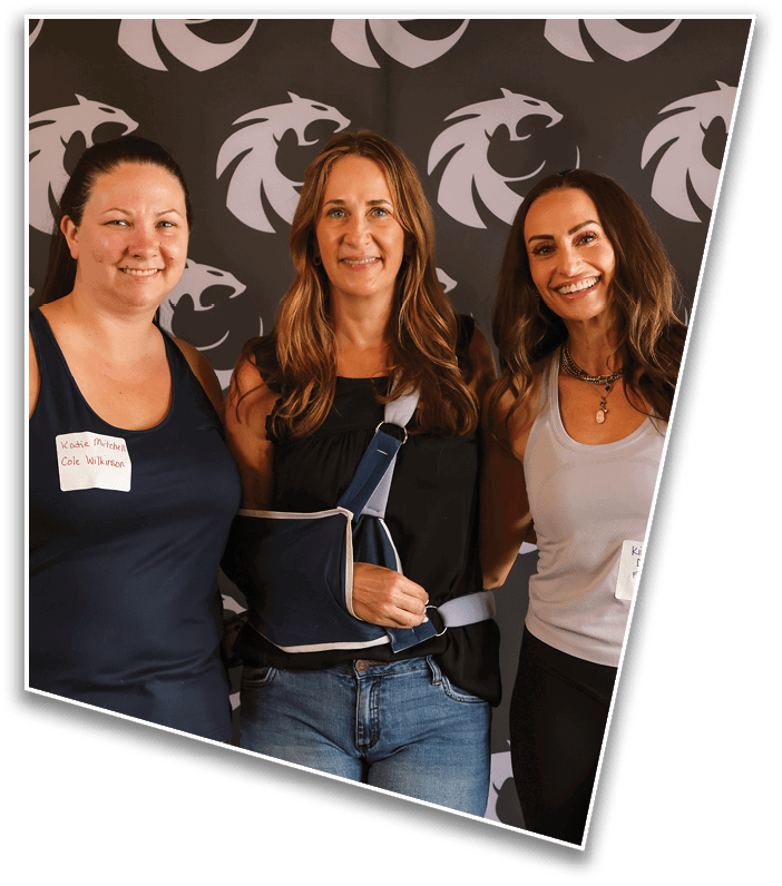Three adults smiling and standing close together in front of a backdrop with a repeating dragon logo, with one person wearing a sling on their arm. Description generated by AI