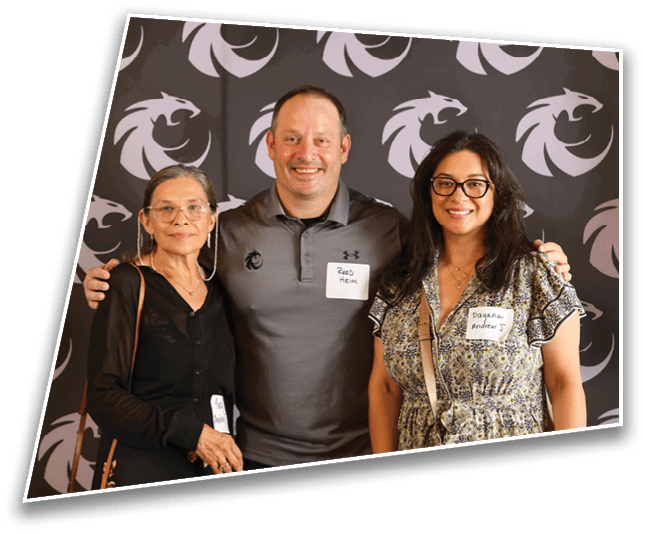 Three people stand closely together smiling in front of a backdrop with a repeating dragon logo, each wearing name tags and casual clothing. Description generated by AI