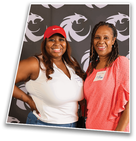 Two adults smiling and posing together in front of a backdrop with a repeating stylized animal logo, one wearing a red cap and white sleeveless top and the other wearing a coral eyelet blouse. Description generated by AI
