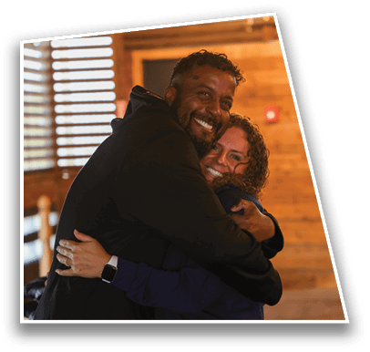 Two adults with curly hair warmly embrace and smile indoors against a wooden wall background. Description generated by AI