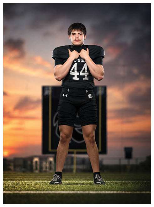 A person wearing a black football uniform with the number 44 stands confidently on a field at sunset with a blurred scoreboard in the background. Description generated by AI