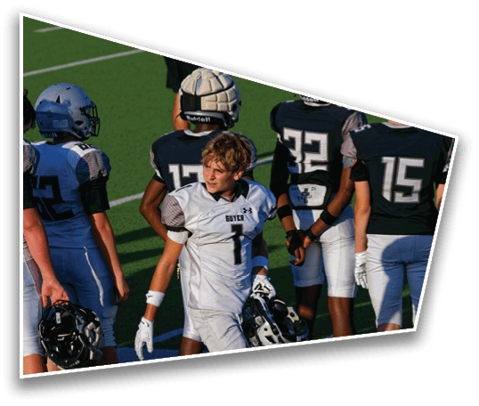 A group of people wearing football uniforms are standing on a field, with one person in a white uniform holding a helmet and looking to the side. Description generated by AI