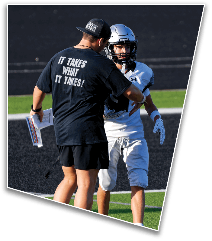 A person wearing a black cap and a black shirt with white text is talking to a person in a white football helmet and uniform against a dark background. Description generated by AI