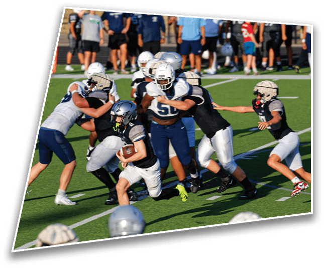 A group of people wearing football helmets and uniforms are actively engaged in a game on a green field, with one person running while holding the ball and others attempting to block or tackle. Description generated by AI