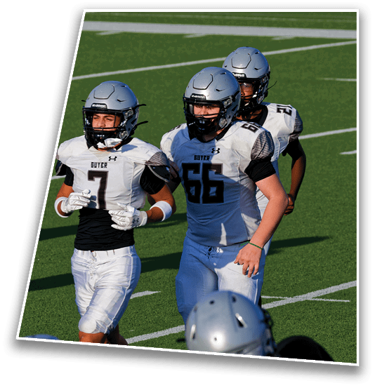 Three people wearing silver football helmets and white jerseys stand together on a green football field. Description generated by AI