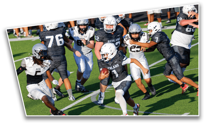 A group of people wearing football helmets and uniforms are actively engaged in a game on a green field with one person holding the ball while others attempt to block or tackle. Description generated by AI