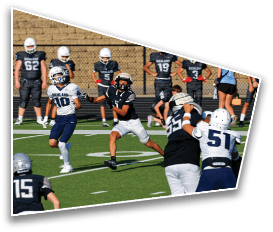 Players in football uniforms are actively engaged in a game on a green field with a brick wall and fence in the background. Description generated by AI