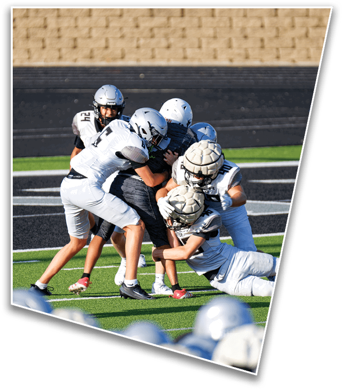 Several people wearing helmets and sports uniforms are engaged in a physical play on a green and black sports field. Description generated by AI