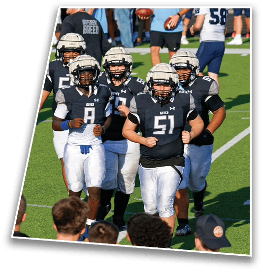 A group of people wearing football helmets and black jerseys are gathered on a field with others in casual sportswear in the background. Description generated by AI