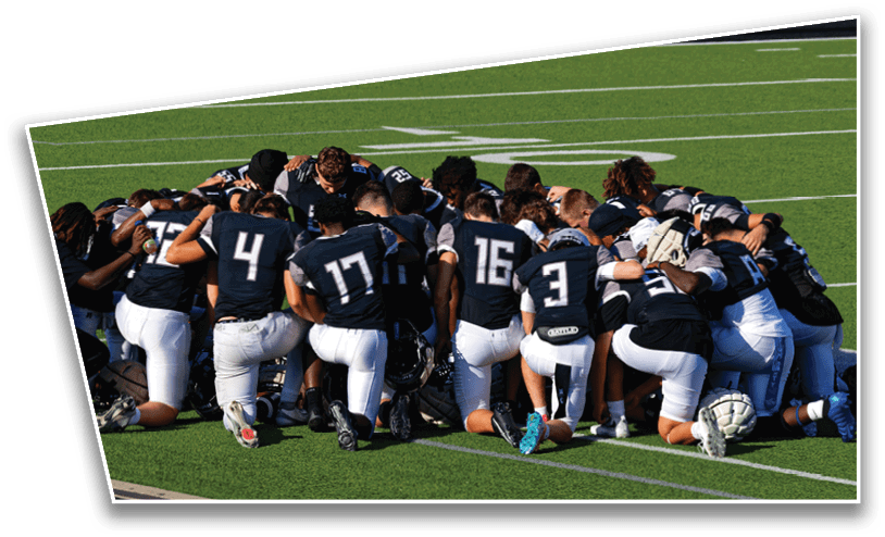 A group of football players in dark jerseys and white pants are kneeling together on a green field in a huddle. Description generated by AI