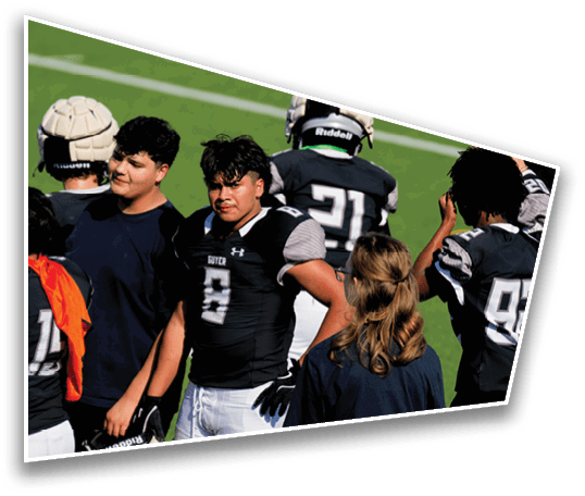 People wearing sports helmets and athletic clothing practice blocking drills on a football field with coaches holding tackling pads. Description generated by AI