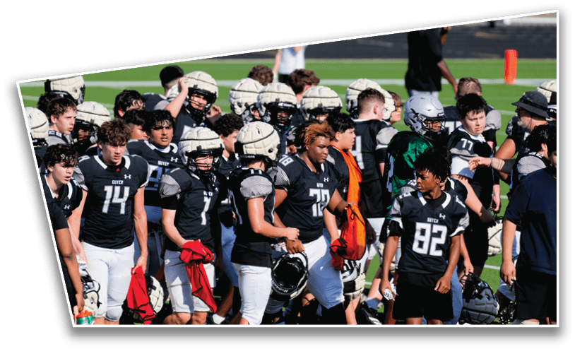 A group of people wearing black and white football uniforms and helmets are gathered on a field, some holding red towels and helmets. Description generated by AI
