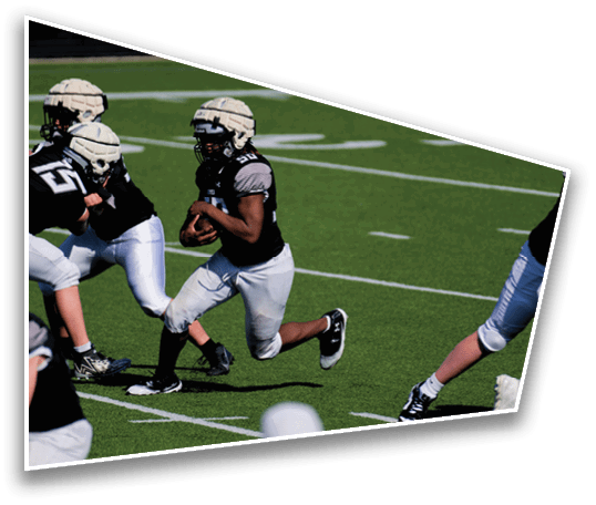 A person wearing a helmet and sports uniform is running with a football on a green field while another person in sports gear is nearby. Description generated by AI