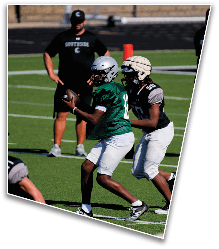 Two people wearing football helmets and uniforms are on a field with one holding a football and the other closely following. Description generated by AI