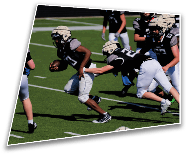 People wearing helmets and football uniforms are playing on a green field with one person running while holding a football and another reaching to grab them. Description generated by AI