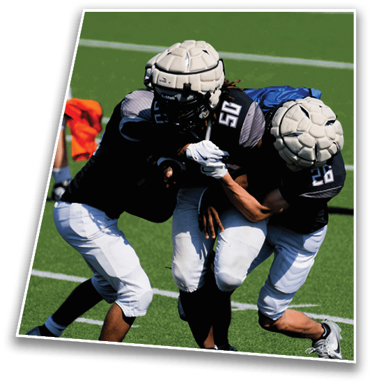 Two people wearing padded helmets and sports uniforms are engaged in a close physical play on a grassy field. Description generated by AI