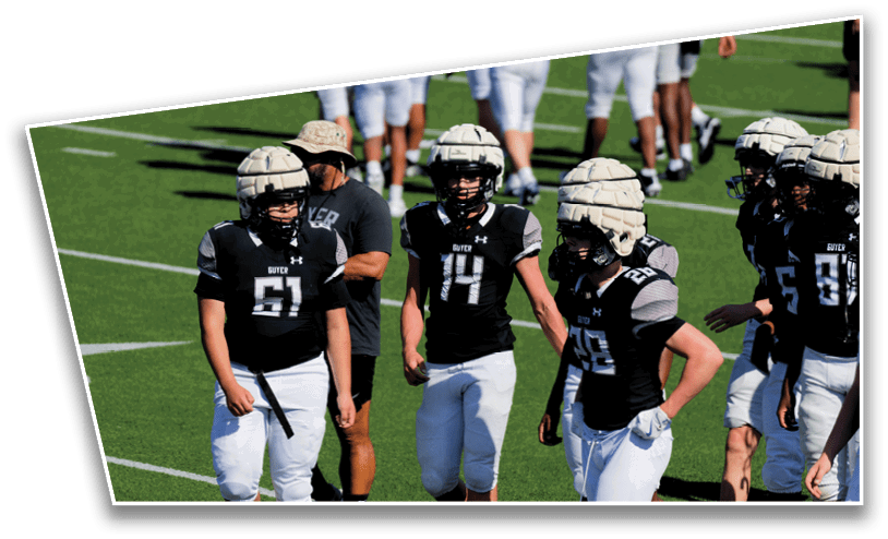 Several people wearing black and white football uniforms and padded helmets stand on a green field with others in the background. Description generated by AI